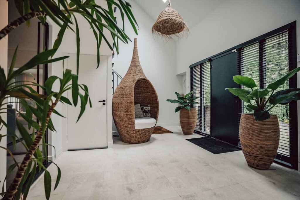 A modern renovated hallway with sleek travertine floors, a minimalist steel door, and a sculptural conic chair. A stylish staircase and lush greenery complete the bright, contemporary design of MYTIPEE.com