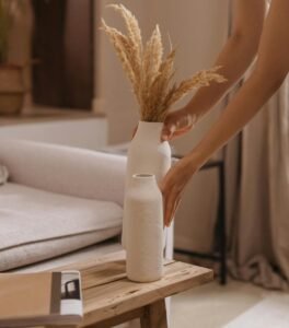 Minimalist hands placing a white ceramic vase with dried pampas grass on a warm oak table — a quiet moment of Japandi-inspired styling.