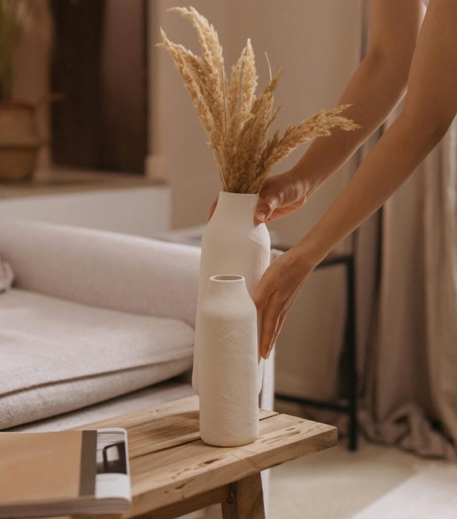 Minimalist hands placing a white ceramic vase with dried pampas grass on a warm oak table — a quiet moment of Japandi-inspired styling.