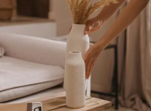 Minimalist hands placing a white ceramic vase with dried pampas grass on a warm oak table — a quiet moment of Japandi-inspired styling.